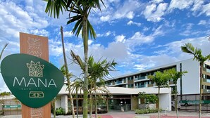 Naamplaatje voor 'MANA Beach Resort' met een modern gebouw op de achtergrond.