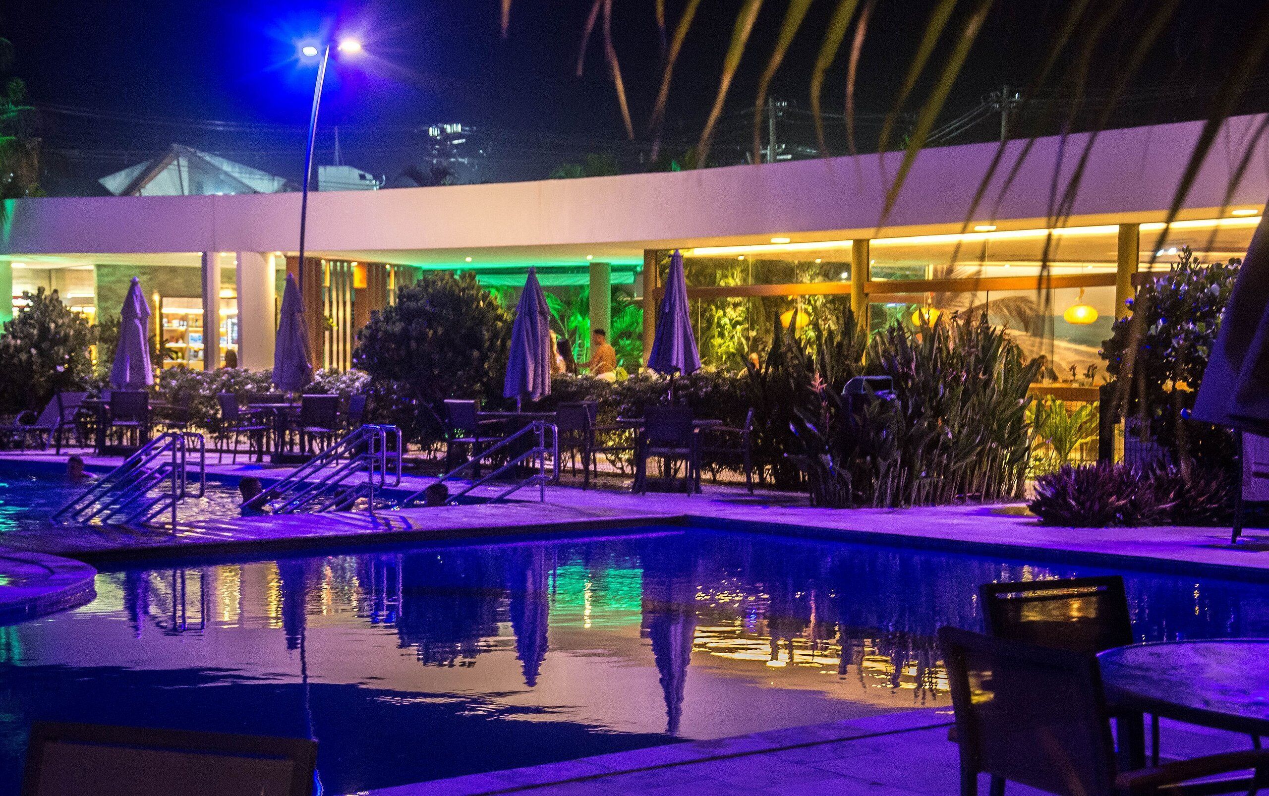 A nighttime pool area with lounge chairs and umbrellas, surrounded by greenery and a building with large windows.