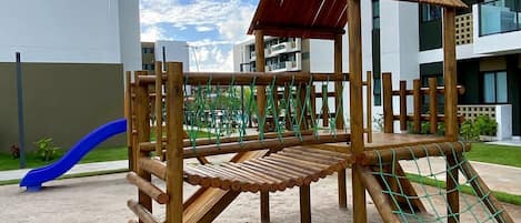 A playground with a slide and climbing structures, set against a modern building with balconies.