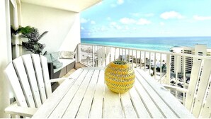 Un balcon avec une chaise blanche, une petite table et un vase décoratif. Le balcon donne sur une plage, avec des bâtiments visibles à l’arrière-plan.