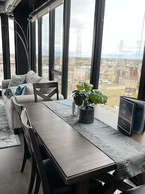 A modern living room with a sofa, dining table, and potted plant. Large windows offer a city view.
