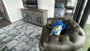 Un salon moderne avec un pouf en cuir, un téléviseur posé sur un meuble, un tapis à motifs et un lit avec une tête de lit.