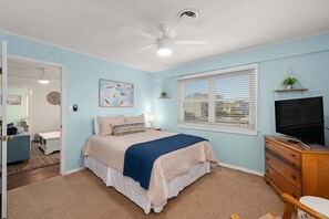 A bedroom with a bed, a TV, a dresser, a window with blinds, and a ceiling fan.