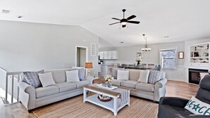 Ein modernes Wohnzimmer mit zwei Sofas, einem Couchtisch und einem schwarzen Ledersessel. Man sieht einen Deckenventilator, einen Kamin und eine Küche im Hintergrund.