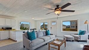 Amplia sala de estar con un sofá gris, una mesa de centro y un ventilador de techo. Hay una zona de cocina con armarios blancos y un microondas. La habitación tiene ventanas amplias que ofrecen vistas del vecindario.