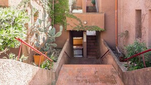 Una escalera conduce a la entrada de un edificio con una reja metálica, rodeada de plantas y un sendero de ladrillos.