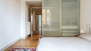 Un dormitori amb un llit, un radiador, una separació de vidre i un passadís que condueix a una altra habitació.