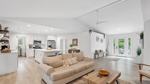 A spacious living area with a beige sofa, wooden coffee table, and a kitchen in the background. - Casa Del Mar - Chic, Large Fenced Yard, Pet Friendly, 0.3 mi to Beach (Folly Beach)