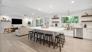 A modern kitchen with a central island, bar stools, a dining area, and a large window. - Casa Del Mar - Chic, Large Fenced Yard, Pet Friendly, 0.3 mi to Beach (Folly Beach)