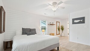 A bedroom with a large bed, a ceiling fan, a window with blinds, a bedside table, and a wall-mounted shelf with a plant. - Casa Del Mar - Chic, Large Fenced Yard, Pet Friendly, 0.3 mi to Beach (Folly Beach)