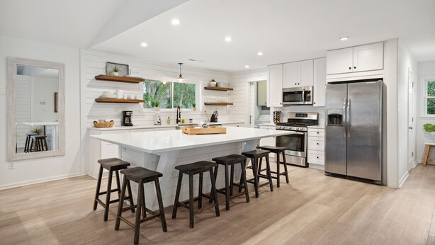 A modern kitchen with white cabinetry, a central island with stools, stainless steel appliances, and wooden flooring. - Casa Del Mar - Chic, Large Fenced Yard, Pet Friendly, 0.3 mi to Beach (Folly Beach)