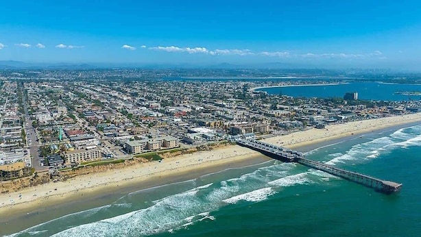 An aerial view of a coastal city with a long pier extending into the ocean, a wide beach, and dense urban development.