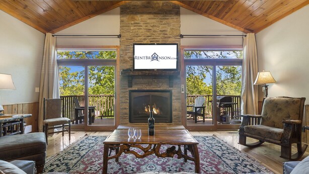 A cozy living room with a fireplace, a television mounted on the wall, and a balcony with outdoor furniture. - Luxurious Adirondack Lodge at Taneycomo with Hot Tub, Stunning Views & Free SDC Tickets (Branson)