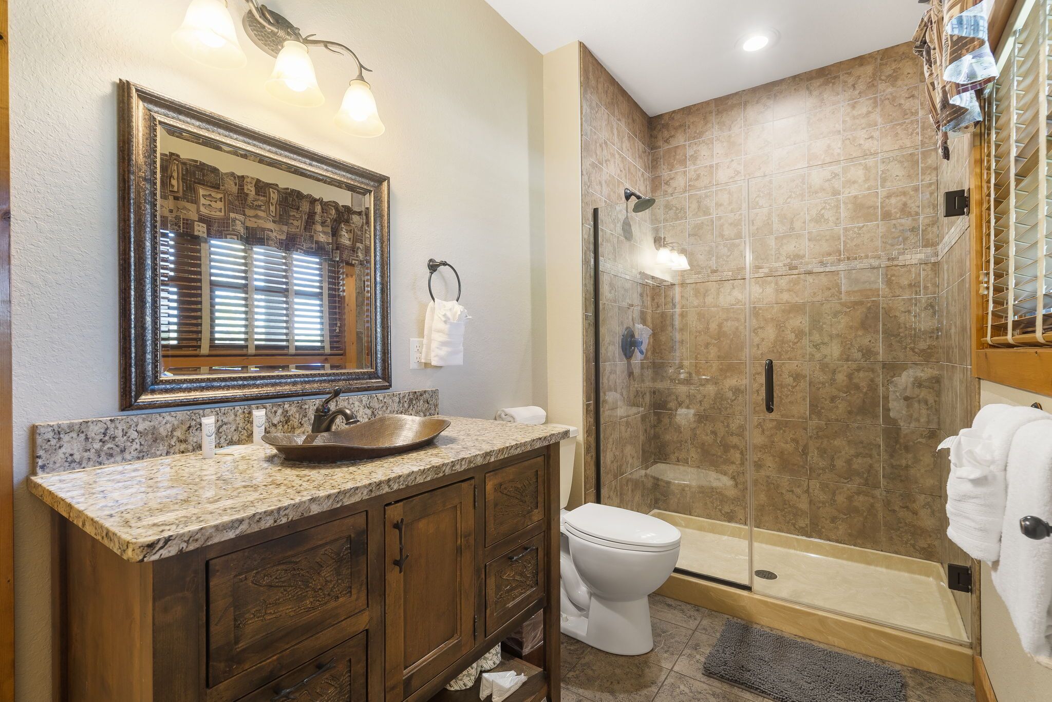 Une salle de bain dotée d’un comptoir en granit, d’un meuble-lavabo en bois, de toilettes blanches, d’une douche à parois de verre et d’un miroir avec un cadre décoratif.