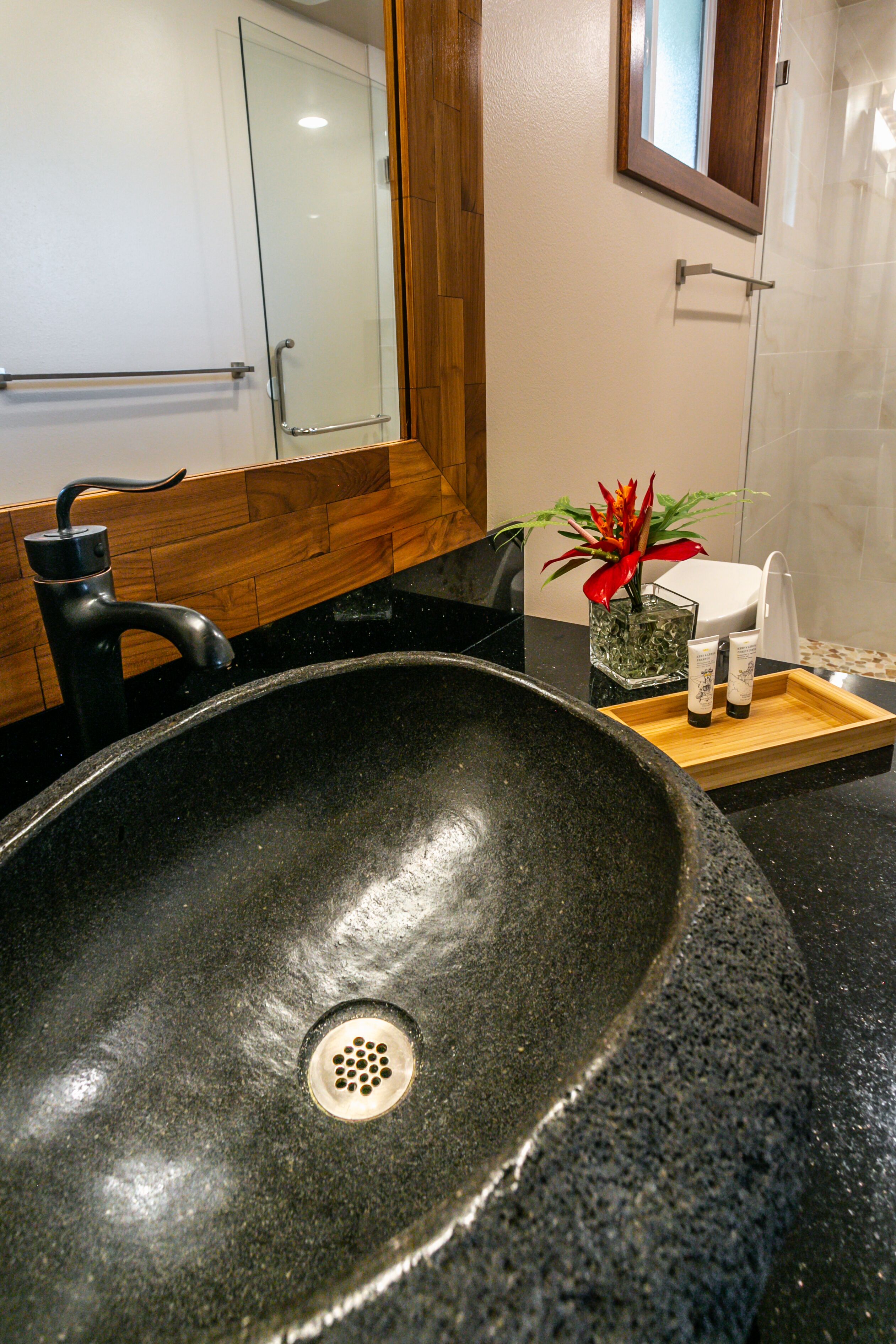 A modern bathroom with a large black sink, a wooden vanity, a glass shower enclosure, and a small plant on the counter.