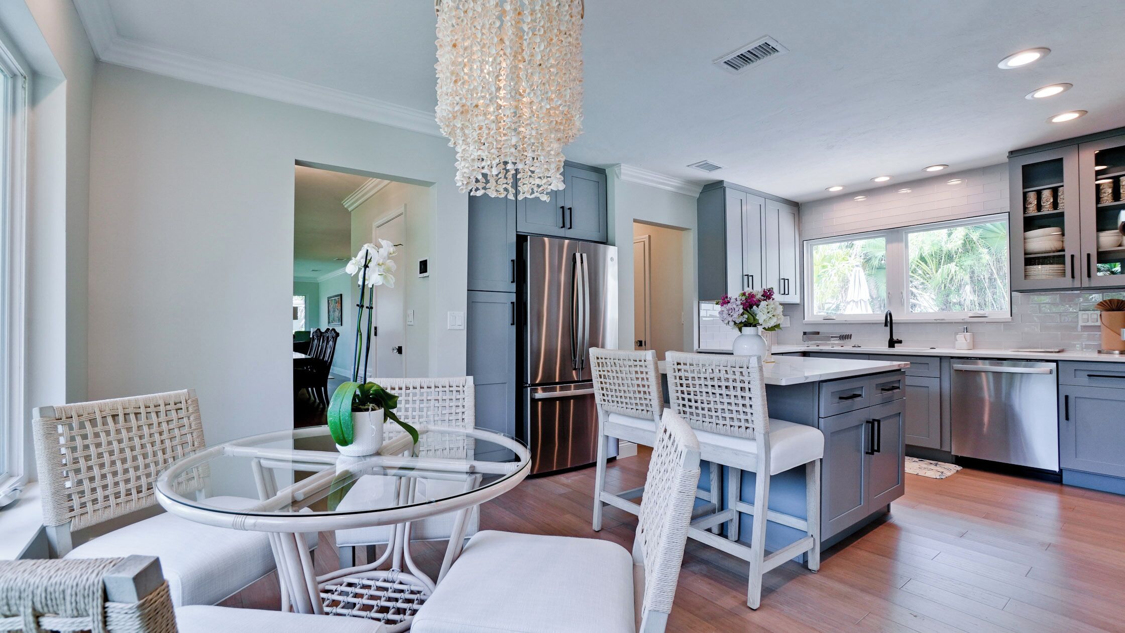 A modern kitchen with stainless steel appliances, a dining area with white wicker chairs, and a large window overlooking greenery.