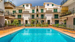 A rectangular swimming pool surrounded by a tiled deck, with a row of multi-story buildings featuring balconies and green shutters in the background. - Suite Home Astrid (Mascali)