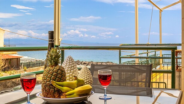 A table with a plate of fruit and two glasses of red wine, overlooking a beach with a clear sky. - Suite Home Astrid (Mascali)