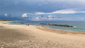 A beach with a rocky breakwater, clear blue water, and distant mountains under a cloudy sky. - Suite Home Astrid (Mascali)