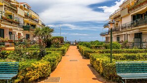 A pathway flanked by neatly trimmed hedges and benches, leading towards a building with balconies and potted plants. - Suite Home Astrid (Mascali)
