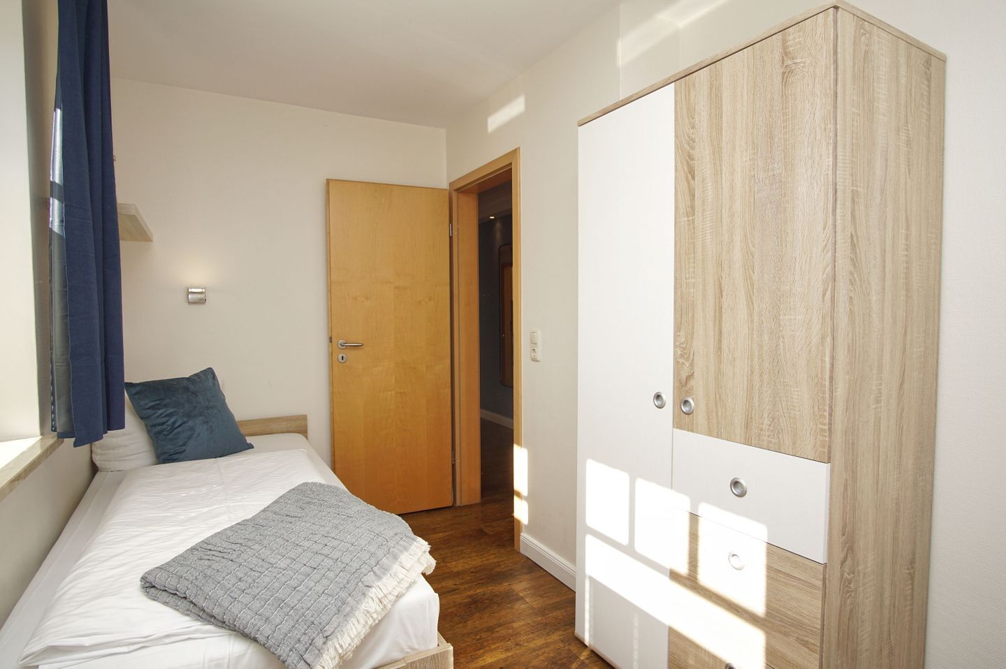 A bedroom with a bed, a wooden wardrobe, and a door leading to another room.