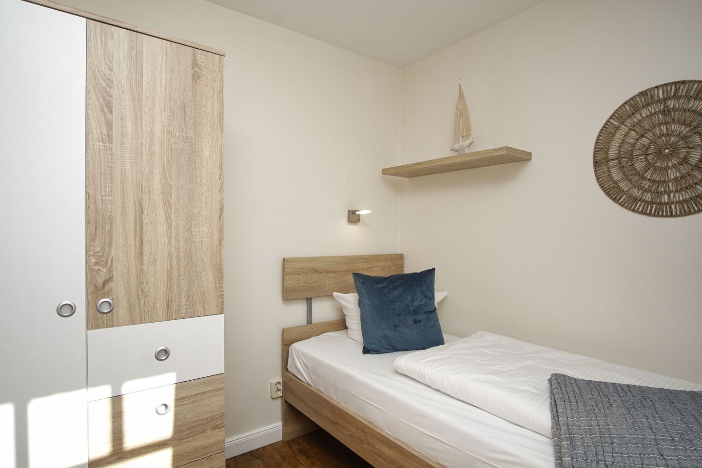 A bedroom with a wooden wardrobe, a bed with white bedding and a blue pillow, a wall-mounted shelf with a decorative item, and a circular woven wall hanging.