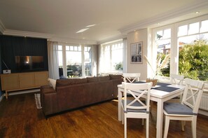 A spacious living room with a large sofa, a dining table with chairs, and a television on a cabinet.