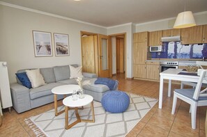 Ein Wohnzimmer mit grauem Sofa, einem runden Couchtisch und einem blauen Pouf. Es gibt eine Küchenzeile mit Holdekor und einem weißen Tisch.
