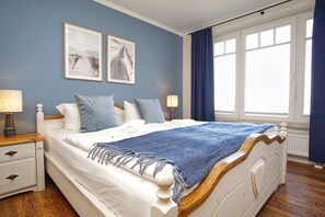 Un lit bien fait, recouvert d’une couverture bleue, avec des oreillers blancs et une tête de lit en bois. Il y a deux tables de chevet avec des lampes et un petit tiroir. Les murs de la pièce sont d’un bleu clair et il y a une grande fenêtre avec des rideaux bleus.