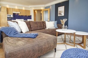 Ein modernes Wohnzimmer mit einem braunen Ecksofa, einem runden Couchtisch und einer Küchenzeile im Hintergrund.