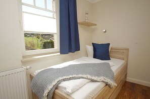 A bedroom with a bed, a window with curtains, a radiator, and a small shelf with a model lighthouse. - Die Ferienwohnung Nr. 04 im Haus am Meer - Haus am Meer (Lohme)