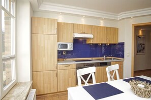 A modern kitchen with wooden cabinets, a microwave, a stove, and a sink. There is a window providing natural light and a table with chairs. - Die Ferienwohnung Nr. 04 im Haus am Meer - Haus am Meer (Lohme)