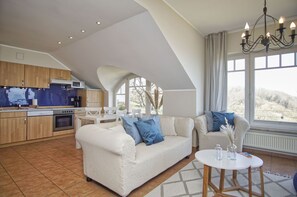 A spacious living area with a sofa and chairs, a dining table, and a kitchen with wooden cabinets and a built-in oven. - Die Ferienwohnung Nr. 09 im Haus am Meer - Haus am Meer (Lohme)