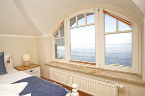 Une chambre avec une grande baie vitrée donnant sur un plan d’eau, un lit, une table de chevet avec une lampe et un radiateur.