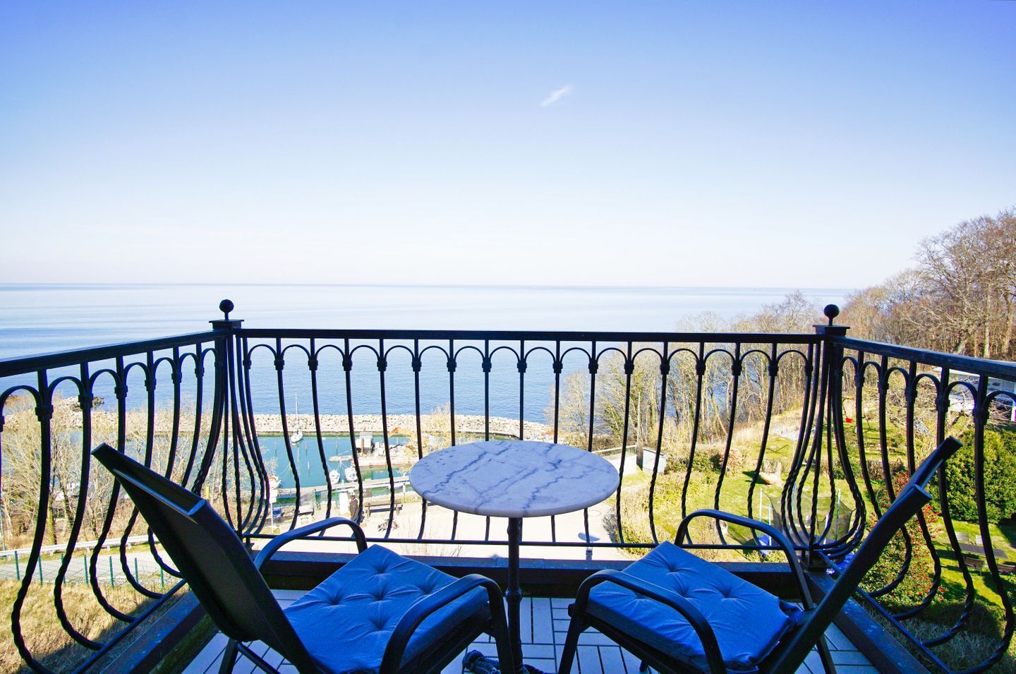 A balcony with a table and two chairs, overlooking a marina with boats and a clear view of the sea.