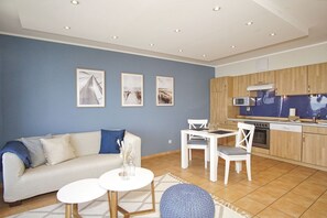 En moderne stue med sofa, to runde borde og et køkken med træskabe og hårde hvidevarer.