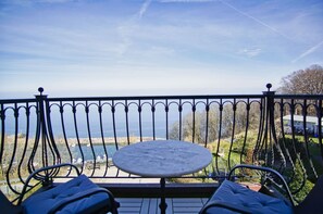 Ein Balkon mit Tisch und Stühlen, der auf ein Gewässer und eine entfernte Küstenlinie blickt.