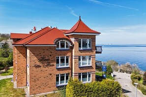 Ein zweistöckiges Backsteingebäude mit rotem Ziegeldach, einem Balkon und Blick auf ein Gewässer.