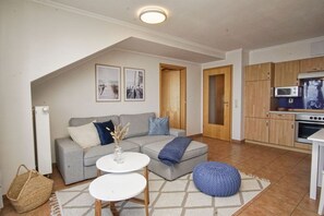 Ein Wohnzimmer mit grauem Sofa, zwei weißen runden Tischen, einem blauen Strickpouf und einer Küche mit Holzschränken und Geräten.