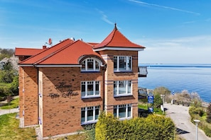 Ein zweistöckiges Backsteingebäude mit rotem Ziegeldach, einem Balkon und Blick auf ein Gewässer.