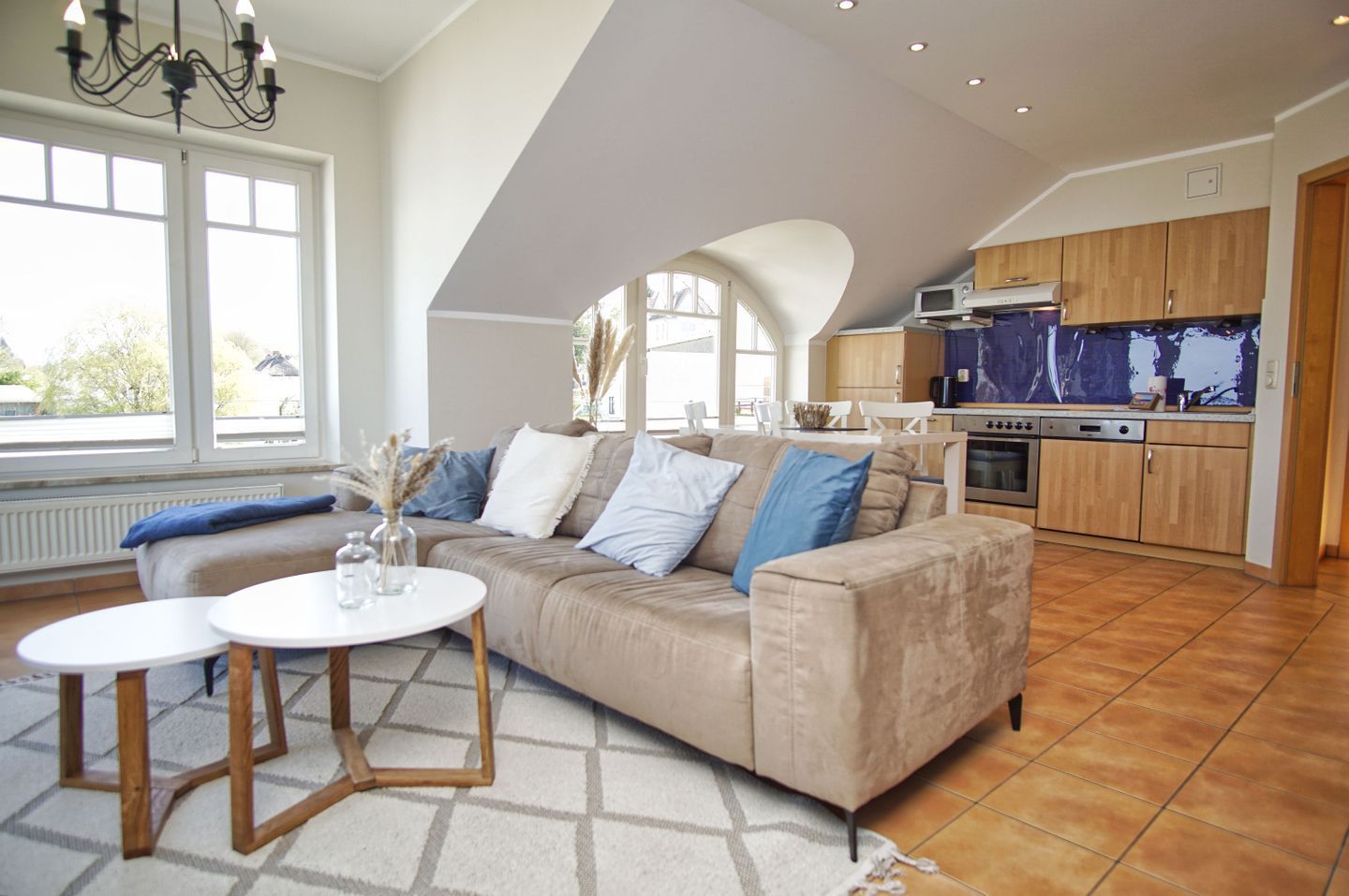 Een moderne woonkamer met een beige bank, twee witte ronde tafels, en een keuken met houten kasten en een groot raam.
