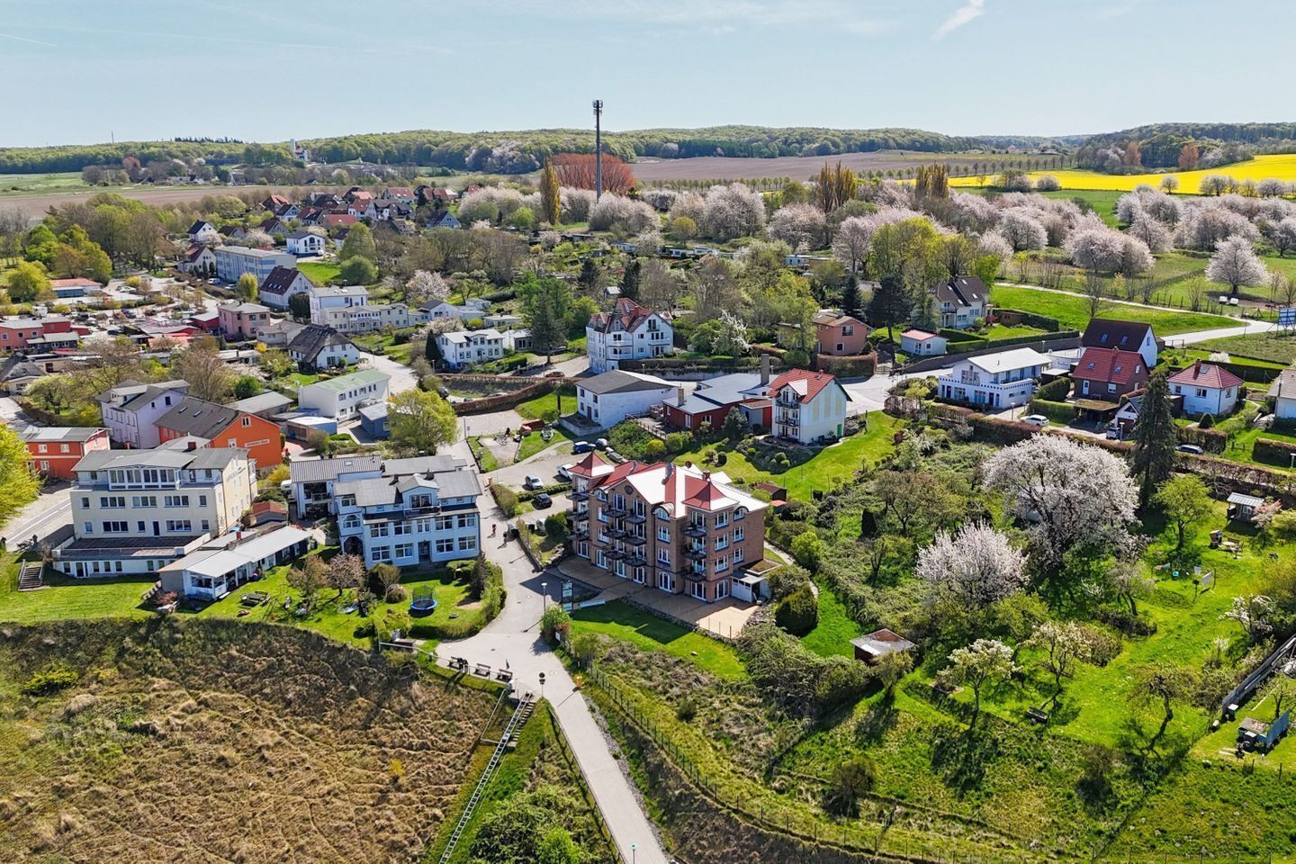 An aerial view of a residential area with houses, a central road, and surrounding greenery.