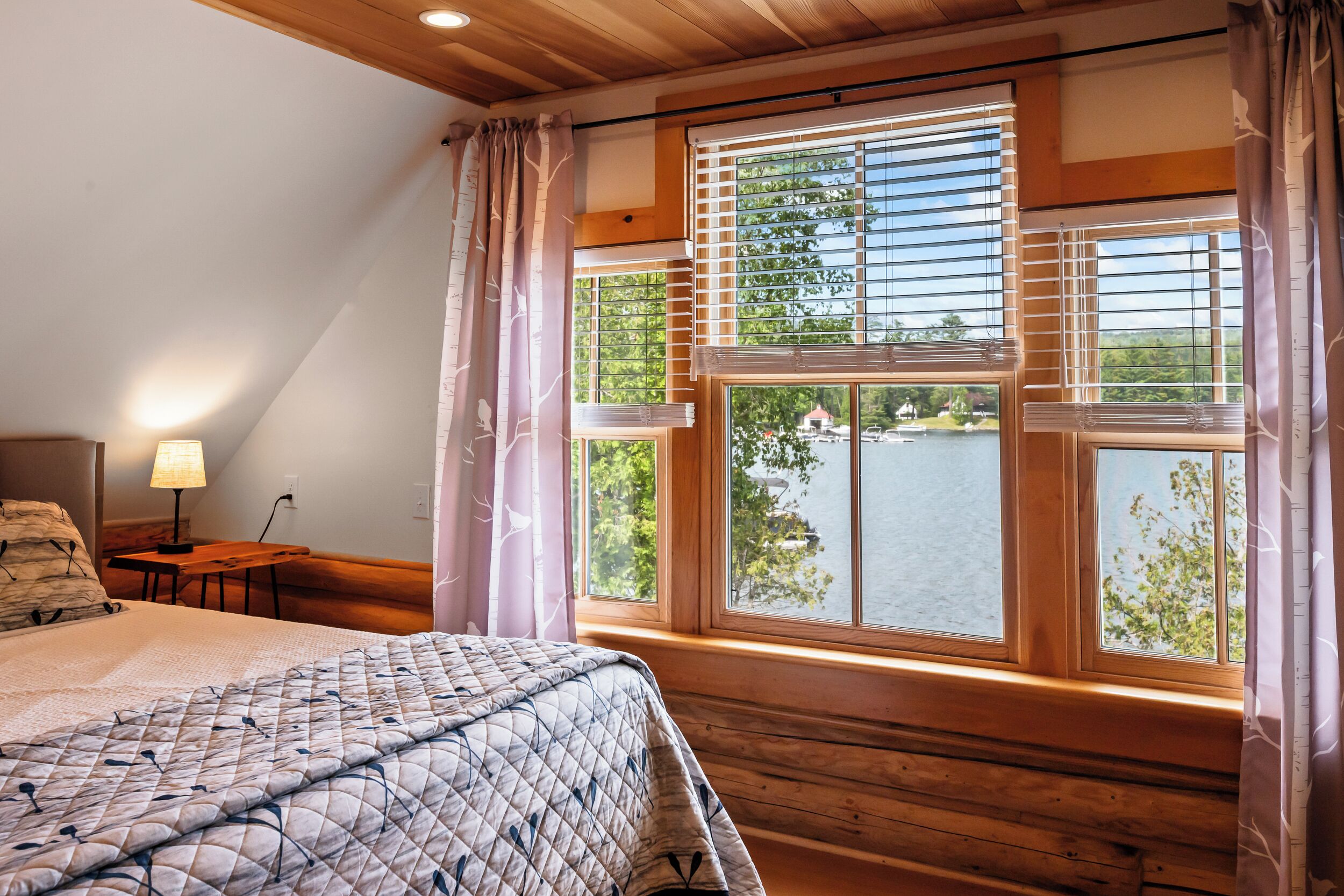 Une chambre à coucher avec un grand lit, une fenêtre avec des stores, une table de chevet et une lampe.