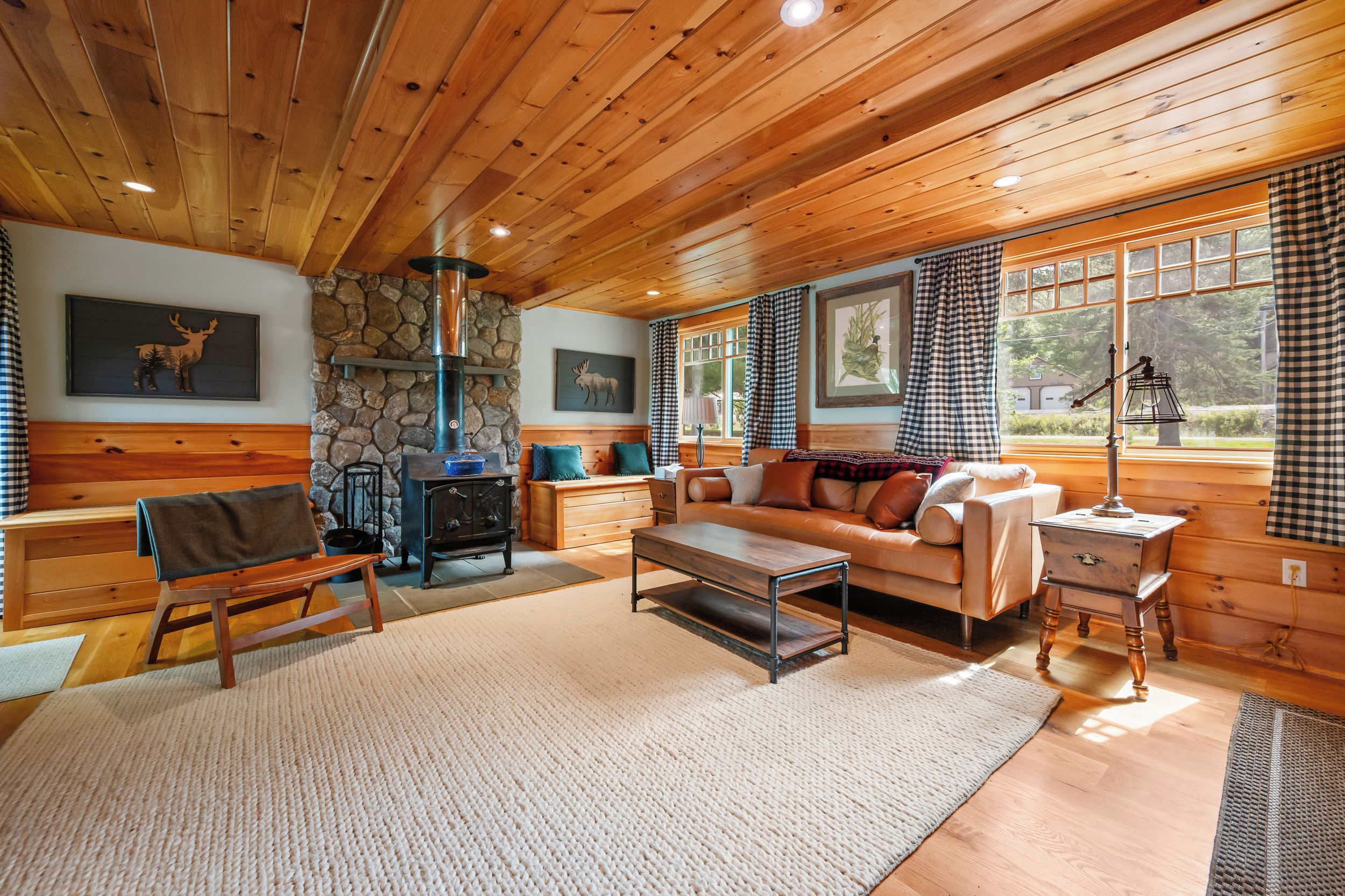 L’intérieur d’un chalet accueillant, avec une cheminée en pierre, des meubles en bois et une grande fenêtre ornée de rideaux à carreaux.
