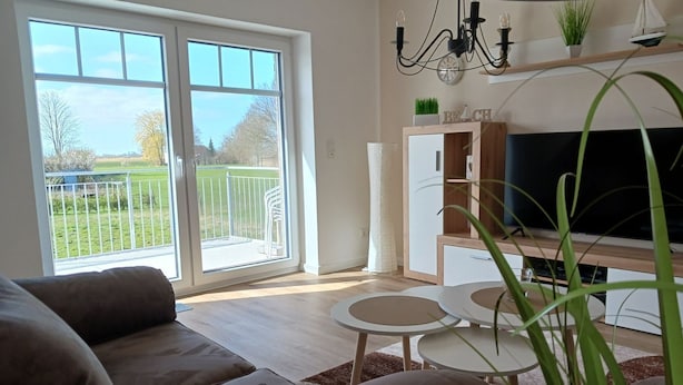 A living room with a sofa, a coffee table, and a TV. There is a sliding glass door leading to a balcony with a view of a green lawn and trees.
