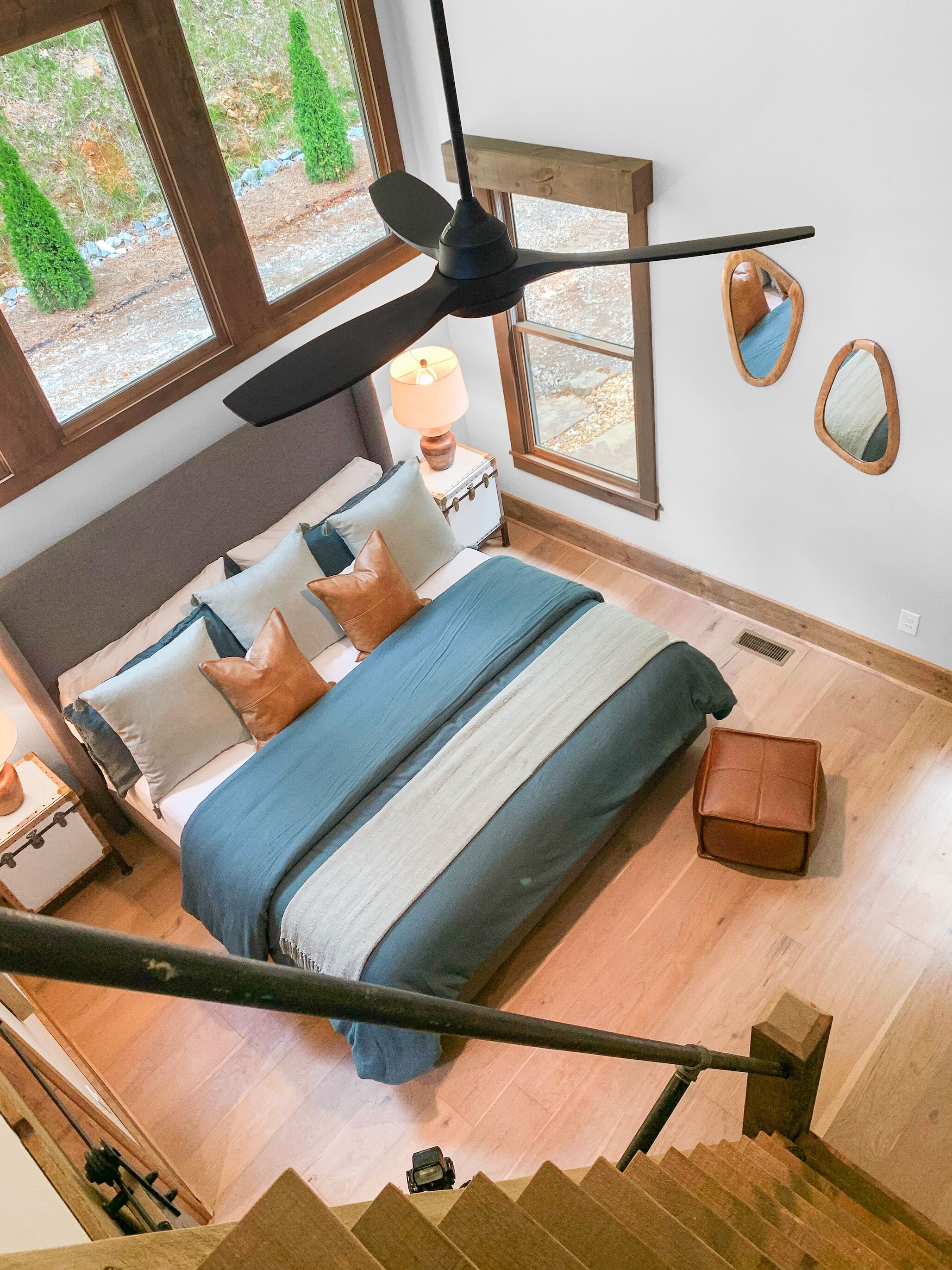 A bedroom with a bed, a ceiling fan, a nightstand, a lamp, and a window with a view of greenery.