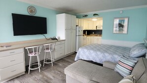 Quarto compacto com cozinha compacta, uma cama com almofadas listradas, um sofá e uma TV fixada na parede.