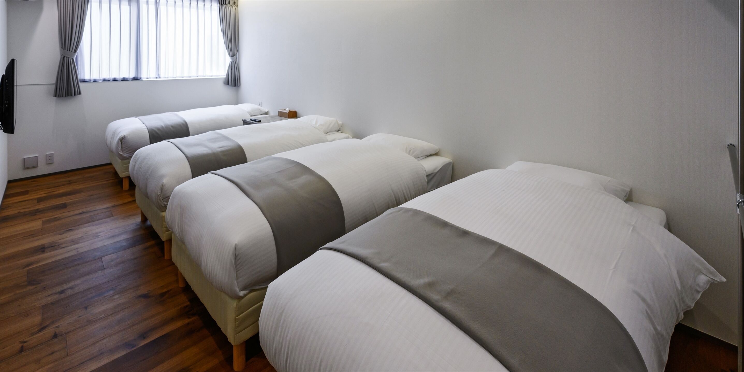 A hotel room with three beds aligned in a row, a wooden floor, a window with curtains, and a flat-screen TV mounted on the wall.