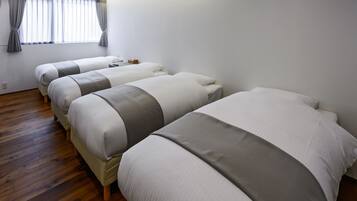A hotel room with three beds aligned in a row, a wooden floor, a window with curtains, and a flat-screen TV mounted on the wall.