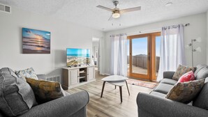 Ein modernes Wohnzimmer mit grauen Sofas, einem Flachbildfernseher, einem Deckenventilator und einem großen Fenster mit Vorhängen.